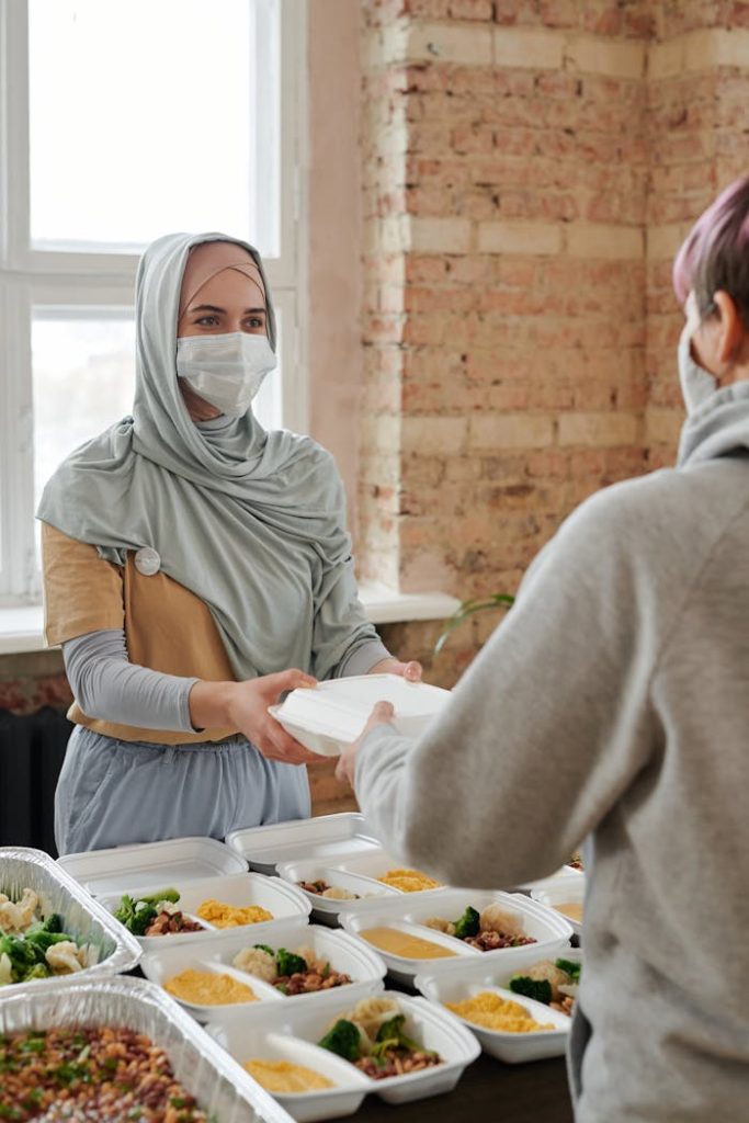a-woman-in-hijab-giving-out-a-packed-food-6995301 A woman wearing a hijab and face mask hands out packed food indoors, promoting charity and safety.